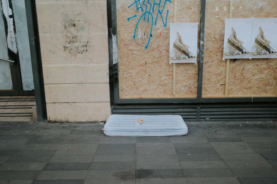 A discarded white foam mattress with a small piece of paper or packaging on top, placed on a paved sidewalk in front of a building with a boarded-up window. The building's exterior wall features a mix